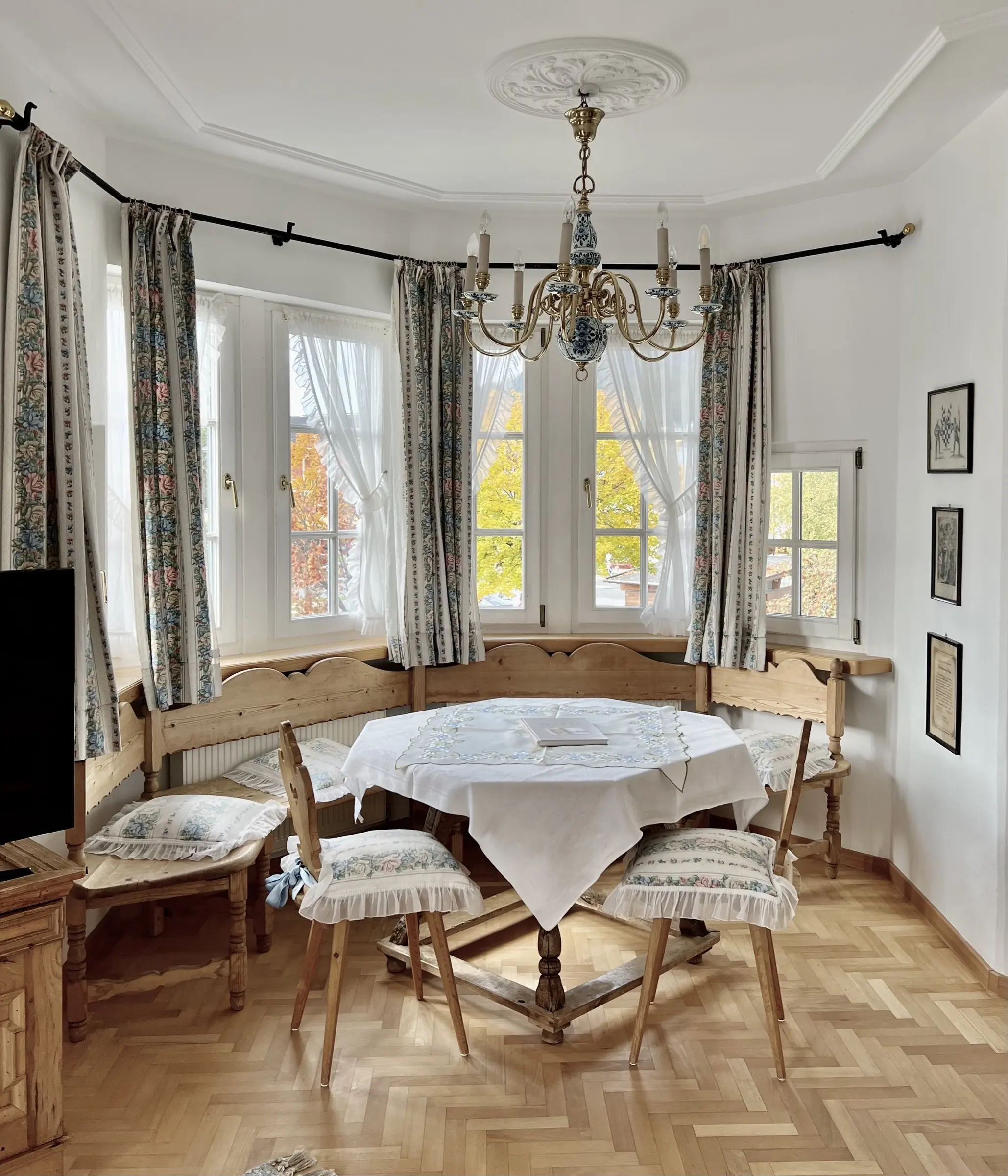 Rincón comedor en el mirador del Apartamento 4: mesa redonda de madera con banco esquinero, lámpara de araña de porcelana azul y blanca, rosetón de estuco y parquet en espiga, árboles otoñales tras las ventanas