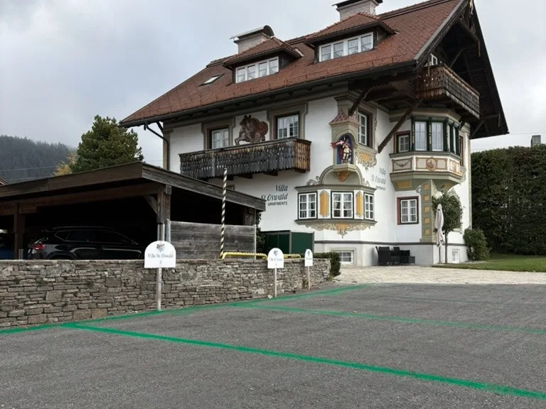 The private parking spaces of Villa St. Oswald, marked in green in front of the stone wall, with the house and its painted facade behind